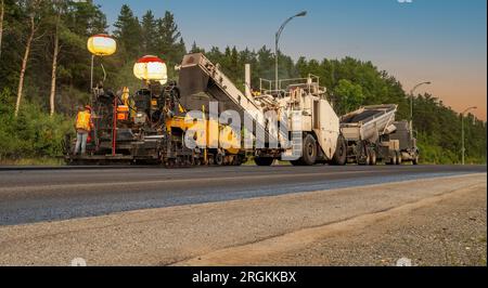 Paving machinery at sunset on a government road, in front of a background of trees and streetlights, vanishing point Stock Photo