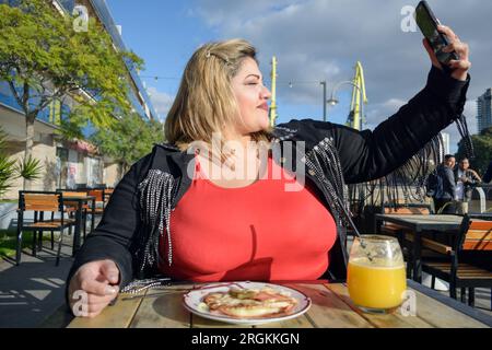 young plus size latin woman sitting outside restaurant at sunset, happy taking selfie photo with her phone and with food served on table, front view, Stock Photo