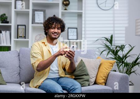 Portrait of young smiling hispanic man at home on sofa smiling and looking at camera, holding phone in hands using app on smartphone. Stock Photo