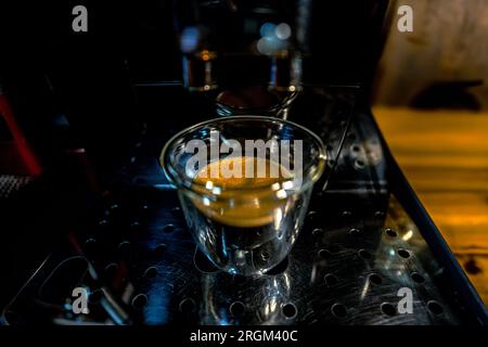 Modern designed glass cup with freshly brewed black coffee with golden foam with small bubbles on top of a barista automatic espresso machine with two Stock Photo