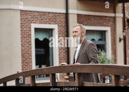 bearded elderly man with grey hair standing on wooden bridge, looking away, thinking, urban backdrop Stock Photo