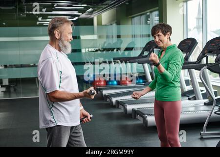 happy woman looking at bearded man working out with dumbbells in gym, active seniors, lifestyle Stock Photo