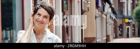 aging population, caucasian, happy, senior woman adjusting short hair, urban backdrop, smile, banner Stock Photo