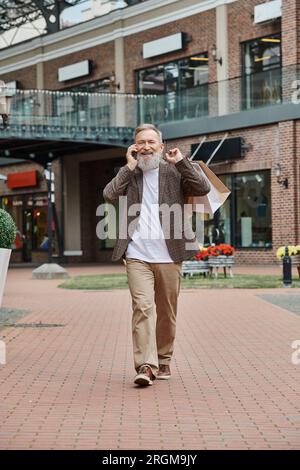happy and elderly man with beard talking on smartphone, holding shopping bags, walking near outlet Stock Photo