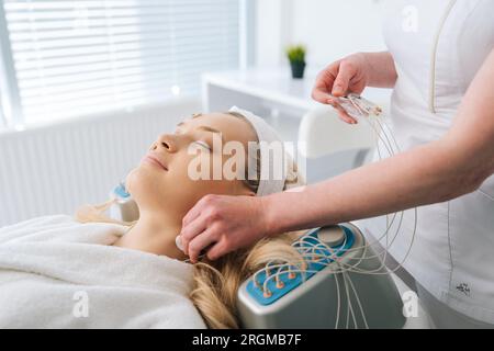 Close-up side view of smiling young woman patient preparing for myostimulation procedure. Beautician putting electrodes of myostimulator Stock Photo