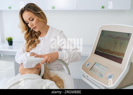 Portrait of Caucasian woman cosmetologist using equipment for ultrasound lifting procedure for blonde female client. Stock Photo