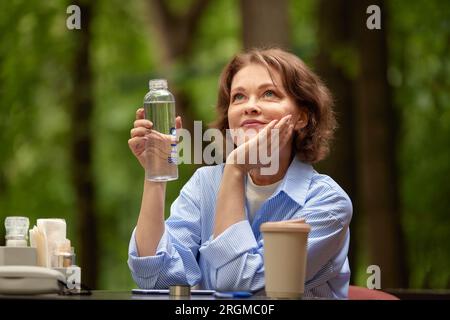 Adorable mature woman sits at the table in outside cafe with bottle of water and paper coffee cup. Senior lady enjoying life on summer terrace. Middle Stock Photo