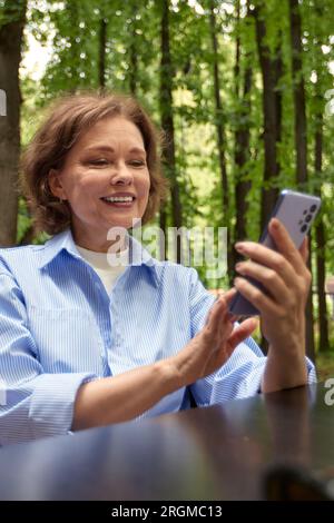 Attractive smiling mature woman at the table in outside cafe. Middle aged brunette female in blue shirt making selfie or video call. Senior lady uses Stock Photo