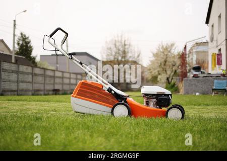 Modern orange-grey electric lawn mower on bright lush green lawn at residential backyard of house. Gardening work tools. Rotary lawn mower machine cut Stock Photo