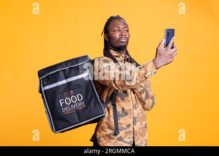 Deliveryman holding phone checking client adreess using takeaway food app before delivery food order. African american restaurant worker carrying thermal backpack standing in studio Stock Photo