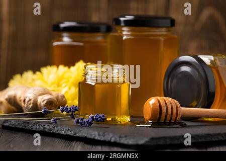 Honey in jars, a wooden dipper lies in a drop of honey, ginger, lavender flowers on a black stone slate plate, close-up. Stock Photo