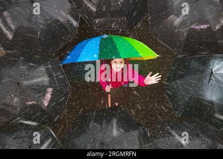 Happy young girl under colorful umbrella in dark crowd in autumn rain. Happiness and optimism concept. Joy and hope in difficult situations. Stock Photo