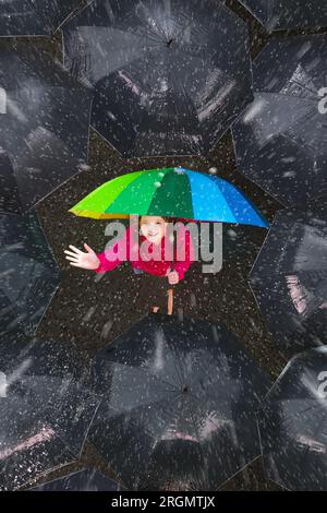 Happy young girl under colorful umbrella in dark crowd in autumn rain. Happiness and optimism concept. Joy and hope in difficult situations. Stock Photo