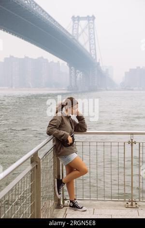 Teen girl posing under Williamsburg bridge in Domino Park, Brooklyn, in a misty and foggy day. Telephoto, vertical portrait. Stock Photo