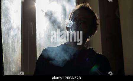 Guy smokes in morning. Man in background of window in smoke. Smoke from cigarettes in room. Silhouette of man in apartment. Stock Photo