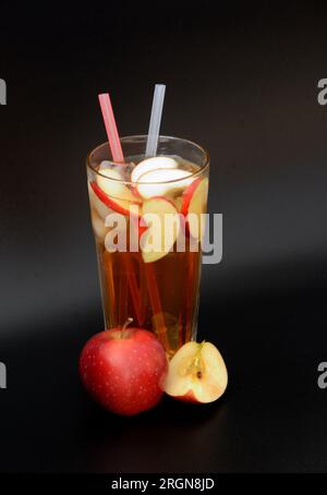 A tall faceted glass of fruit juice with ice and straws on a black background, next to it are slices of a ripe red apple. Vertical arrangement. Stock Photo