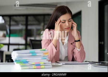 A stressed and unhappy Asian businesswoman sits at her desk with an unhappy face, suffering from headaches, tired of overwork, and feeling overwhelmed Stock Photo
