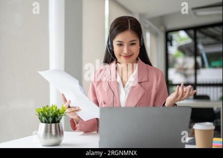 A beautiful Asian female customer service operator or call-centre agent in headset is having a conversation with a customer over the phone. Stock Photo
