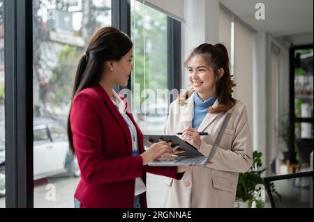 Two beautiful and happy Asian businesswomen or female marketing workers are talking and discussing work in an office corridor. Stock Photo