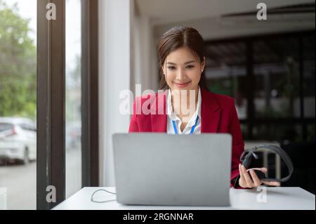 A gorgeous, confident Asian female call centre operator or customer support operator is managing her work on her laptop, working in her office. Stock Photo