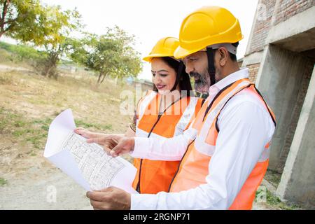 Two Indian male and female civil engineers or architect wearing helmet and vest holding paperwork blueprint at construction site discussing real estat Stock Photo