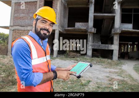 Young Indian male civil engineer or architect wearing helmet and vest holding digital tablet blueprint at construction site. Stock Photo