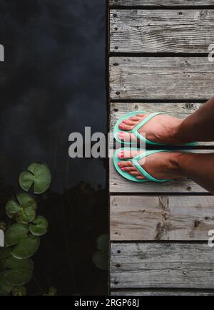 Overhead of woman's feet in sandals on worn wooden dock on lake. Stock Photo