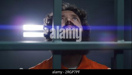 Male criminal in orange uniform holds metal bars with handcuffed hands, stands in prison cell. Prisoner serves imprisonment term for crime in jail. Tired inmate in correctional facility. Rack focus. Stock Photo