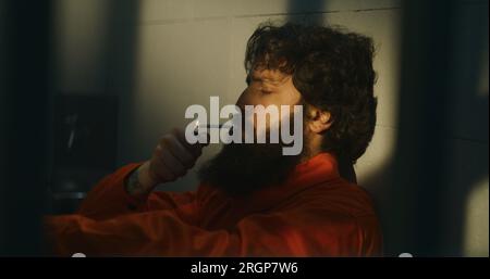 Thoughtful male prisoner in orange uniform sits on the bed, lights cigarette with lighter and smokes in prison cell. Criminal, inmate serves imprisonment term for crime in jail. Detention center. Stock Photo