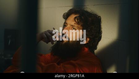 Thoughtful male prisoner in orange uniform sits on the bed, lights ...