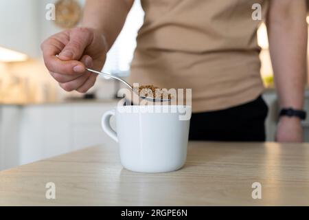 Woman pouring instant coffee granules into a white cup with a spoon. Girl with one spoonful of granulated coffee beans in a kitchen. Stock Photo