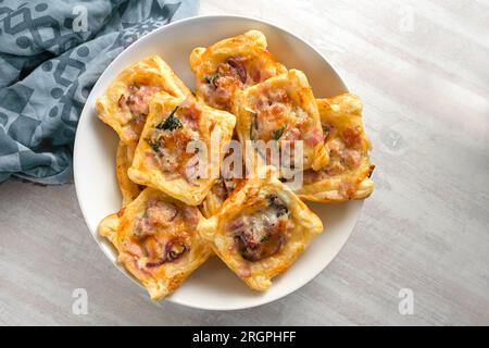 Baked puff pastry squares with ham, onion, herbs and cheese in a bowl, finger food snack for a warm or cold party buffet, high angle view from above, Stock Photo