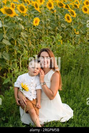Family concept. A beautiful woman with a little boy are sitting on the grass in a field with yellow sunflowers. Mother gently hugs her son. Stock Photo