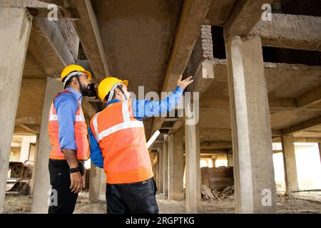 Two Indian female civil engineers or architect wearing helmet and vest holding paperwork blueprint doing inspection of construction site, Real estate. Stock Photo