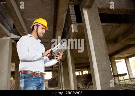 Young handsome Indian male civil engineer or architect wearing helmet holding digital tablet blueprint at construction site. Low angle. Stock Photo