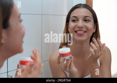 Close-up of relaxed young Brazilian woman applies moisturizer cream on her face after washing in bathroom. Enjoys beauty treatments. Uses cosmetic pro Stock Photo