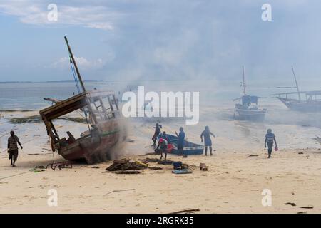 African fishermen set fire under their traditional wooden Dhow boat on the beach to protect it from pests and woodworm and repair boat.Zanzibar,Tanzan Stock Photo