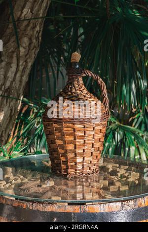 Large Vintage Wicker Wrapped Wine Bottle. Old Demijohn with Cork in sunset rays on a glass table with wine corks under glass. Stock Photo