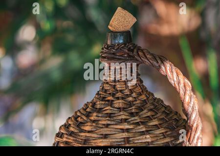 Large Vintage Wicker Wrapped Wine Bottle. Old Demijohn with Cork in sunset rays on a glass table with wine corks under glass. Stock Photo