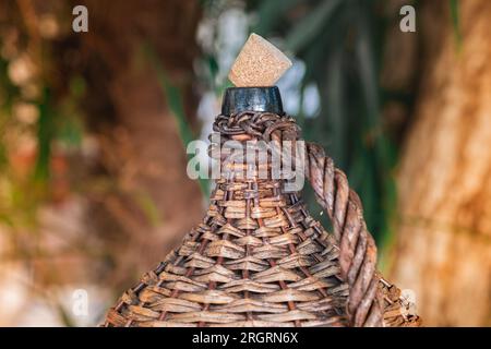 Large Vintage Wicker Wrapped Wine Bottle. Old Demijohn with Cork in sunset rays on a glass table with wine corks under glass. Stock Photo