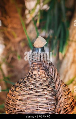 Large Vintage Wicker Wrapped Wine Bottle. Old Demijohn with Cork in sunset rays on a glass table with wine corks under glass. Stock Photo