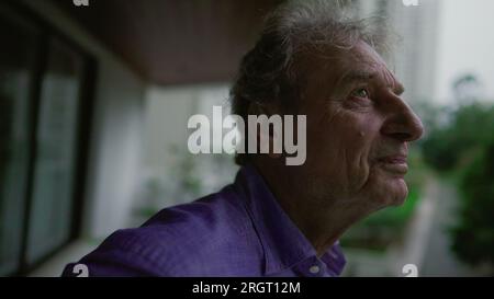 Happy contemplative senior man standing at apartment balcony looking outside in the evening. Thoughtful expression of a older male person in 70s laugh Stock Photo