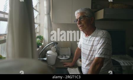 Thoughtful Senior Man Gazing Out of Kitchen Window, Leaning on Counter. Retired older person thinking while staring out window Stock Photo