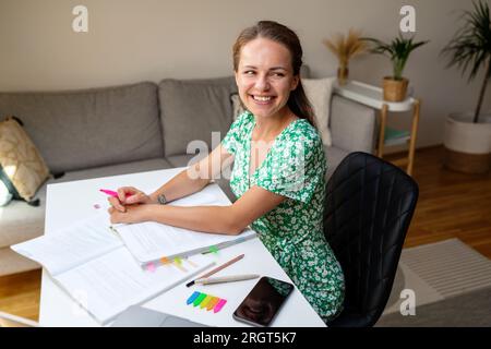 young adult student brunette woman wearing summer dress sits at the desk at home doing a homework and smiling. Stock Photo