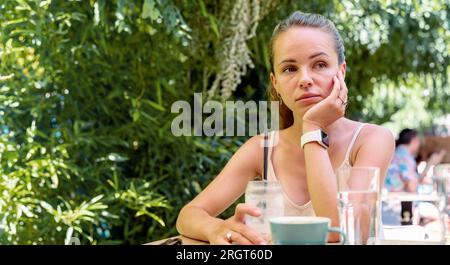 Sad tired urban woman sitting at the table in cafe in summer, mental illness, apathy, loneliness. Stock Photo