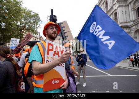 London, UK. 11th Aug, 2023. A junior doctor waves a flag and holds a placard expressing his opinion during a demonstration outside Downing Street. Junior doctor members from the British Medical Association (BMA) continue to take 96-hour walkout monthly a result of dispute over pay and working conditions with the UK Government. The strikes affected tens of thousands of hospital appointments and elective surgeries and starting from 7 am on 11th August to 7 am on 15th August 2023. Credit: SOPA Images Limited/Alamy Live News Stock Photo