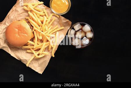Cheeseburger and french fries on brown paper and wooden table, top view. Burger, french fries, cola with ice in a glass and mustard. Fast food Stock Photo