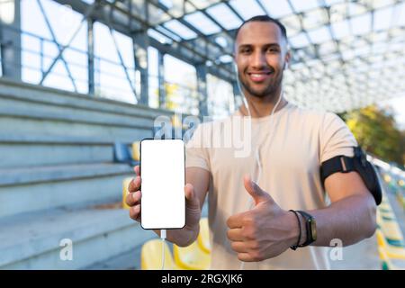 Young hispanic sportsman smiling and looking at camera, man in stadium showing white phone screen, while jogging and doing active exercise. Stock Photo