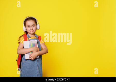 Beautiful little kid girl wearing wireless headphones, carrying backpack, smiles cutely, looking at camera, isolated on orange studio background. Prep Stock Photo