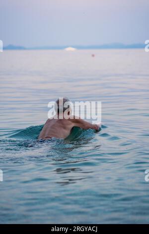 Aged man swims in the Adriatic sea. Gray haired man swimming in tranquise blue water of sea an sunset time. Stock Photo